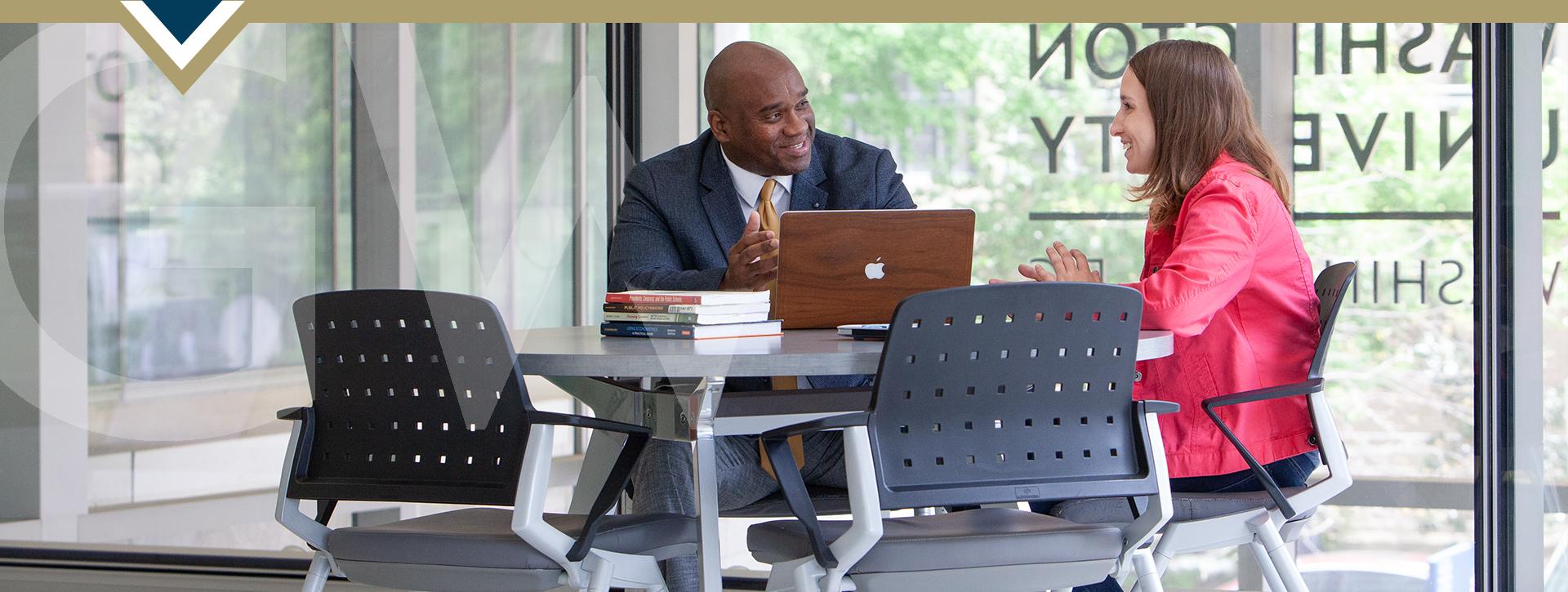 two professionals sit at table with laptop open between; GW logo on window