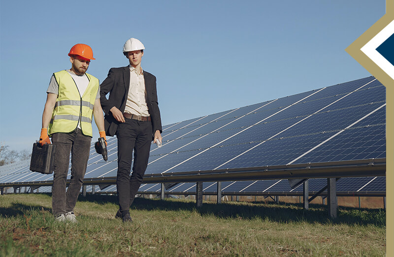two males walking beside a large line of solar panels; one man wearing a suit, the other wearing a work vest and hard hat