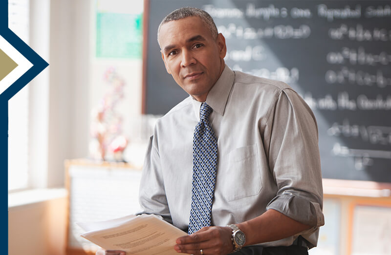 faculty member sits on desk in front of chalkboard in classroom