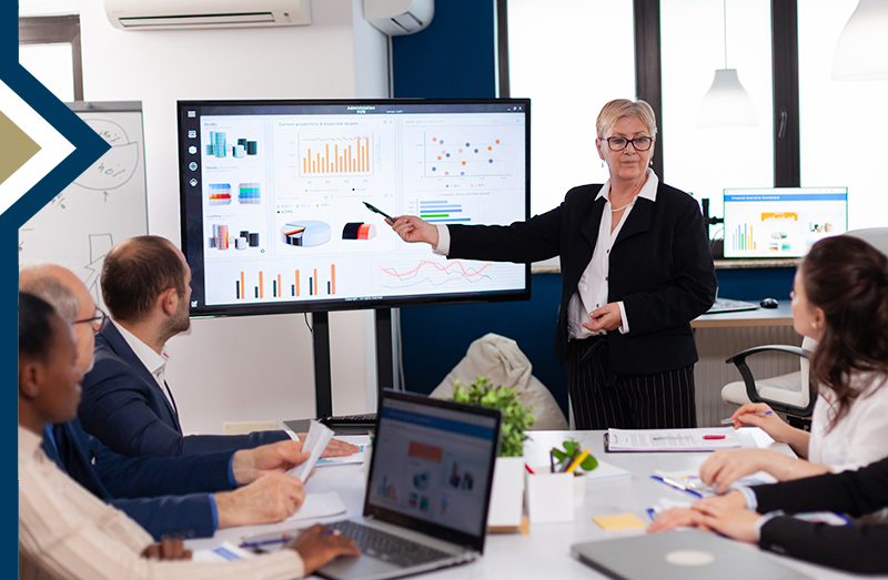 professional points to data on digital screen to group sitting at conference table