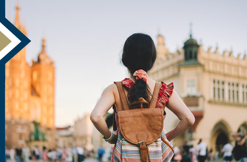 female wearing backpack with back to camera, peering at buildings abroad
