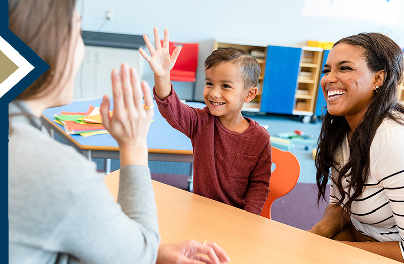 teacher reaches to give preschool student seated across from her a high five, second professional female sitting next to him smiles