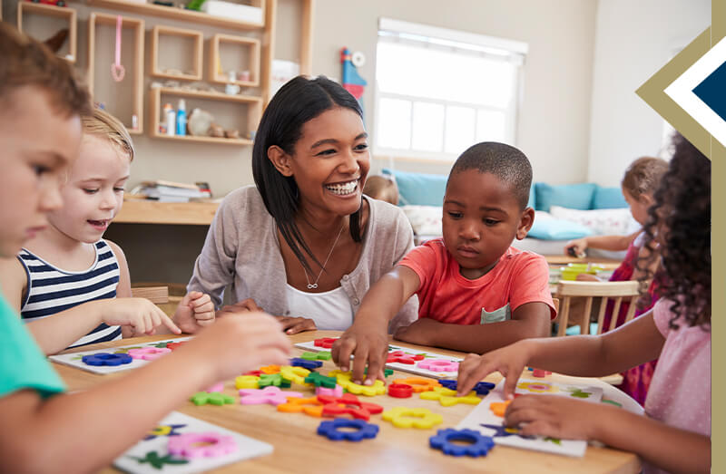 educator/counselor smiles at young children playing with gears on table in classroom