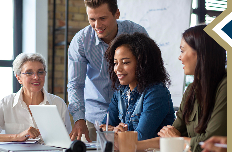 Professional male leans over to assist colleague sitting at table with laptop, two other colleagues are seated next to her - Image by Freepik