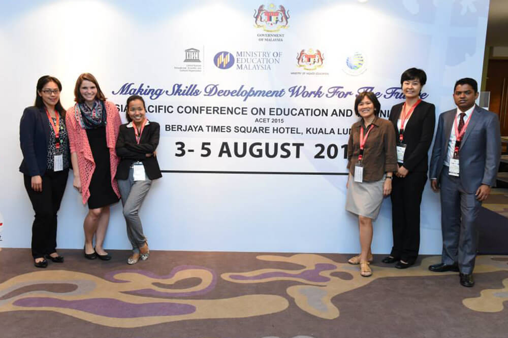 Maggie Appel-Schumacher poses with group of UNESCO staff/fellows at in front of conference signage in Bangkok