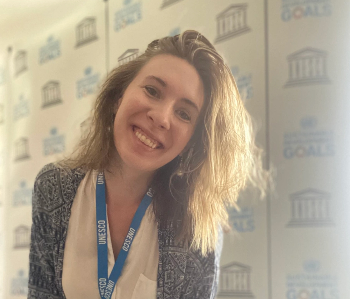 Hayley smiles at the camera wearing a UNESCO lanyard in front of a UNESCO backdrop on her first day in the office