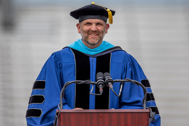 Dr. Mark Blanchard wearing regalia speaks at graduation ceremony