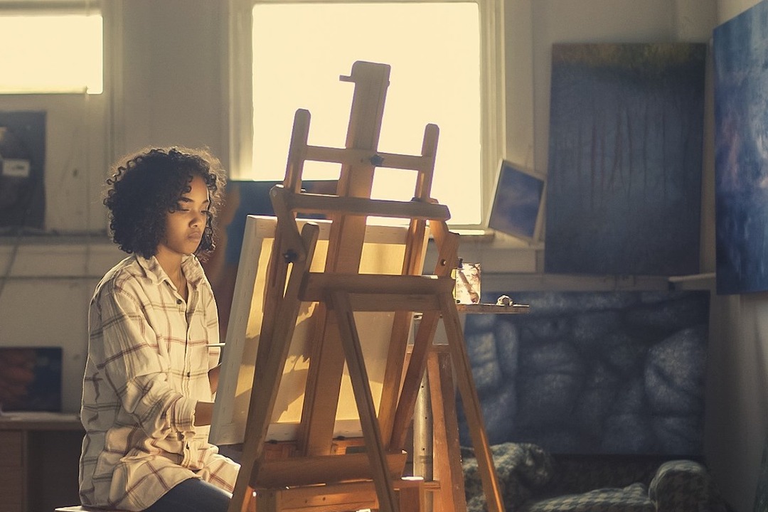 female artist sits behind easel painting, outside light casts a glow over her and her canvas