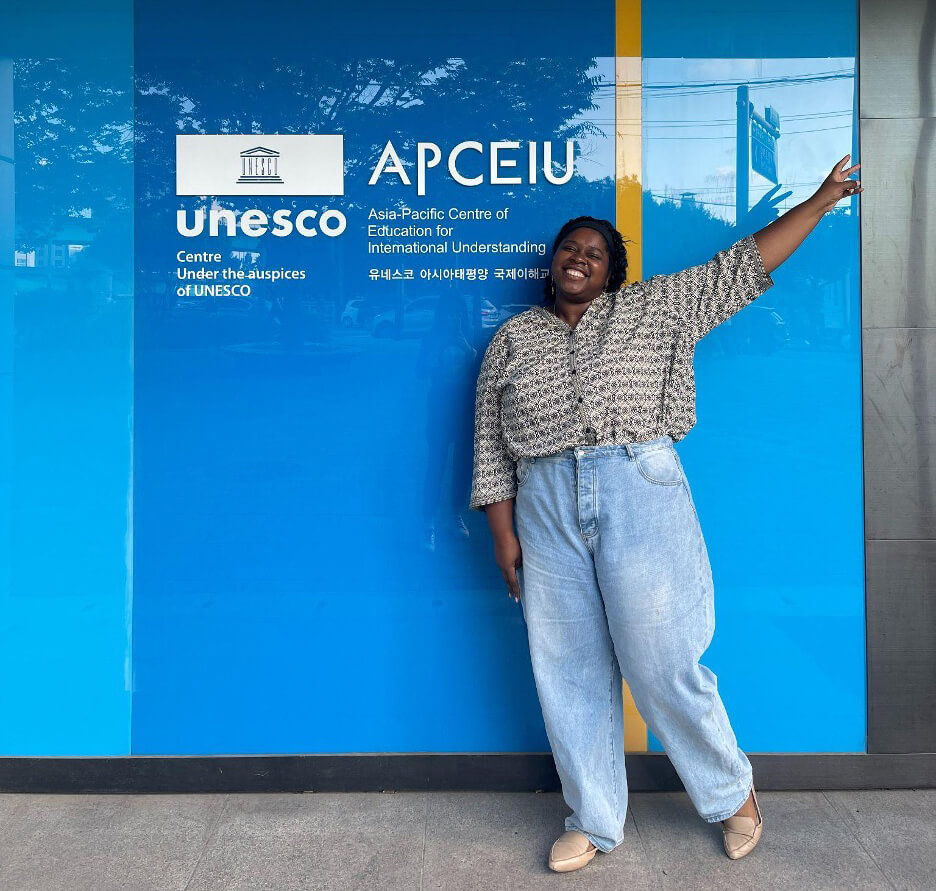 Micha standing and making a peace sign in front of APCEIU logo 
