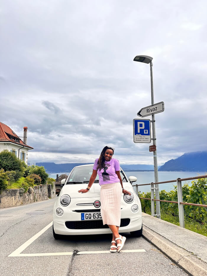 The author, Oummou Diallo, standing in front of a small white Fiat Cinquecento rental car, smiling broadly with mountains and the lake visible in the background.