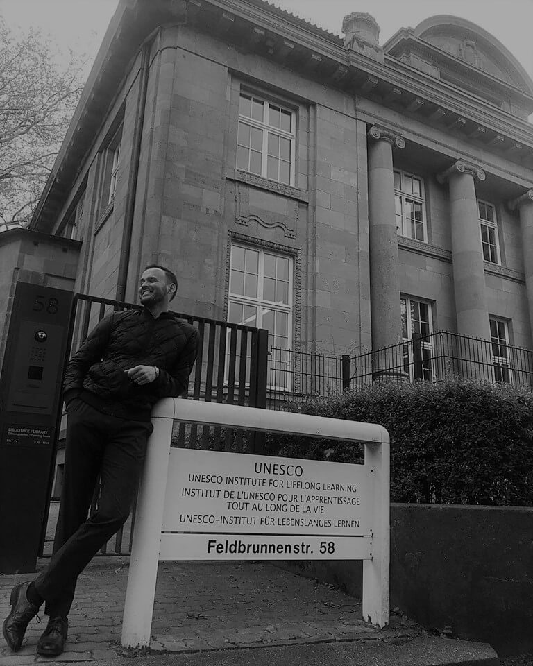 black and white photo of Kyle Hall posing by UNESCO UILL sign in front of the building