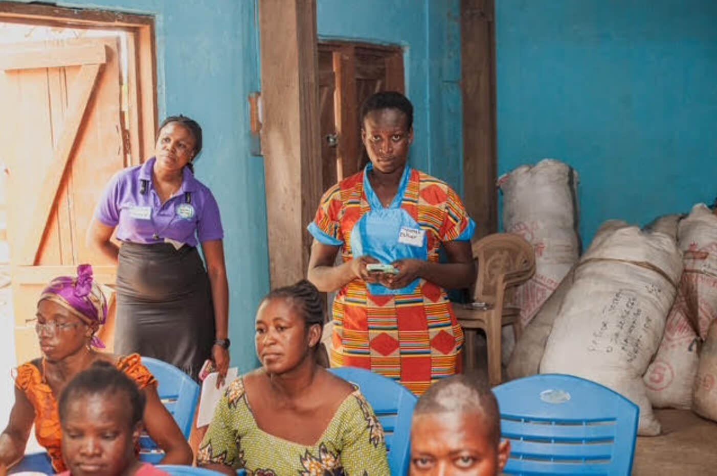 two females guests of IIES event stand in the back of the room, a few women sit before them; large bags lean against the back wall, the door to the room stands open with sun pouring in