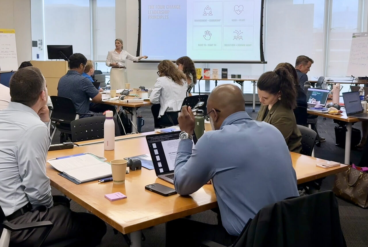 Laurin Parthemos points to the projector screen, course participants seated around the room at square tables, laptops open in front of them