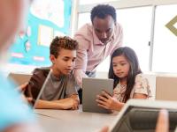 teacher leans down to assist elementary students using tablet