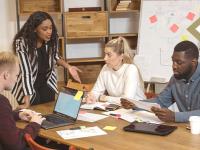 group of four young professionals discuss data on laptop and whiteboard in conference room