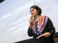 Ina Gjikondi gives a presentation holding microphone as she speaks, presentation on large screen behind her