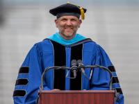 Dr. Mark Blanchard wearing regalia speaks at graduation ceremony
