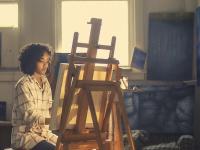 female artist sits behind easel painting, outside light casts a glow over her and her canvas