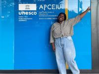Micha standing and making a peace sign in front of APCEIU logo 