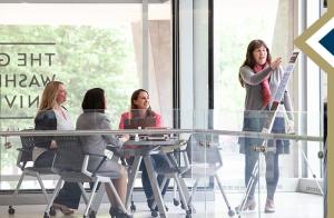 GSEHD faculty member presents information on a board on an easel while three GSEHD students sit at a table and listen
