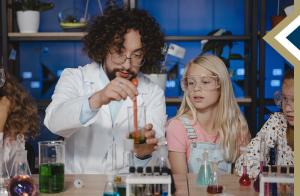 male teacher in white lab coat conducts experiment with colored liquids and beakers while elementary students watch