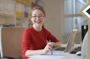 young professional female smiles at camera, looking away from the laptop on her desk
