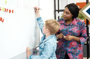teacher watches as an elementary student writes on the white board