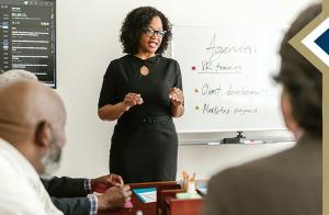 professional female stands in front of white board presenting information to a group of colleagues