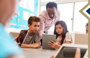 teacher leans down to assist elementary students using tablet