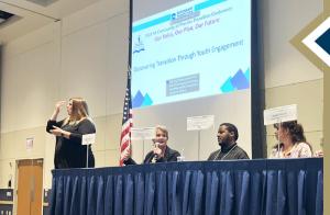 Students participate in panel conversation at conference; one person speaking by podium, others seated behind table; PowerPoint presentation in the background