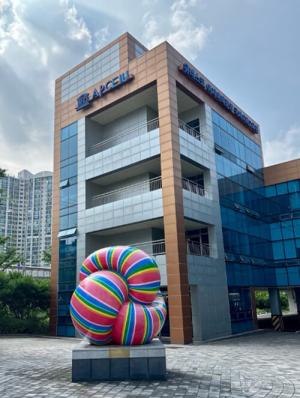 An image of a four story building with “APCEIU” written on the side. There is a colorful art installment in front of the building that resembles a knot or piece of candy.