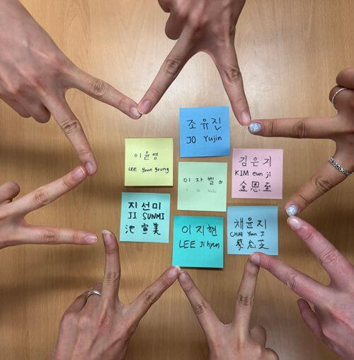 A picture of seven people’s hands all creating a star shape around sticky-notes that have their names written in Korean, English, and Chinese