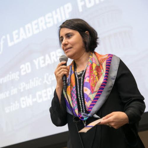 Ina Gjikondi gives a presentation holding microphone as she speaks, presentation on large screen behind her