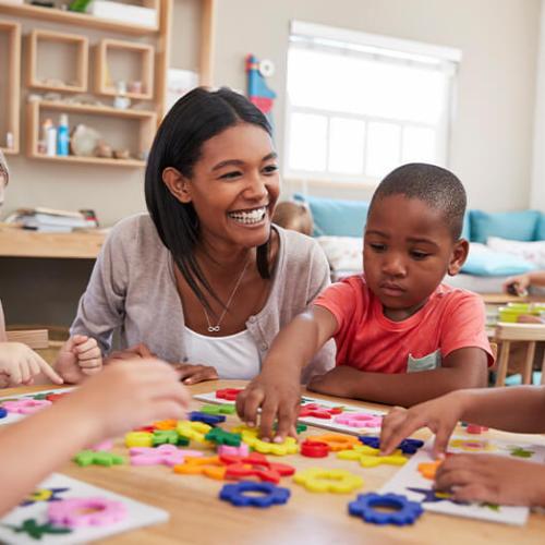 educator/counselor smiles at young children playing with gears on table in classroom
