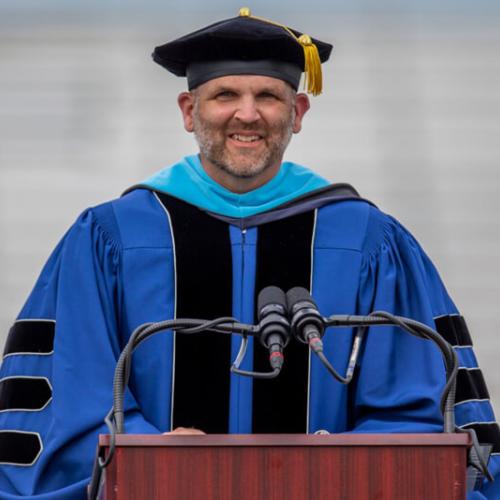 Dr. Mark Blanchard wearing regalia speaks at graduation ceremony