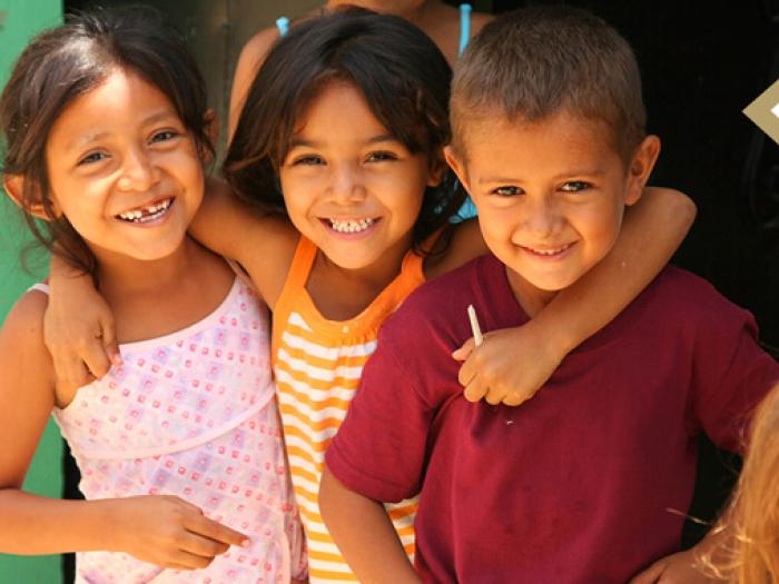 image of three diverse young children smiling at camera, child in middle has her arm around her 2 classmates