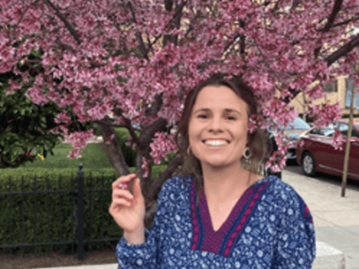 Mara Moettus smiles for photo in front of cherry tree, holds cherry blossom in her fingers
