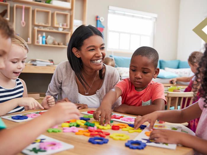 educator/counselor smiles at young children playing with gears on table in classroom