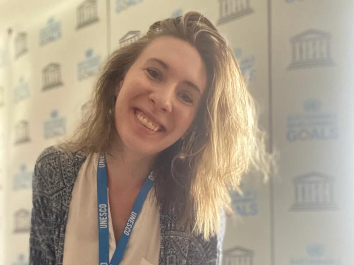 Hayley smiles at the camera wearing a UNESCO lanyard in front of a UNESCO backdrop on her first day in the office