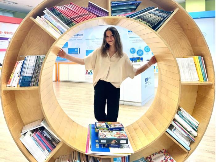 Kseniia poses behind a large round bookshelf, designed to represent the UN Sustainable Development Goals logo