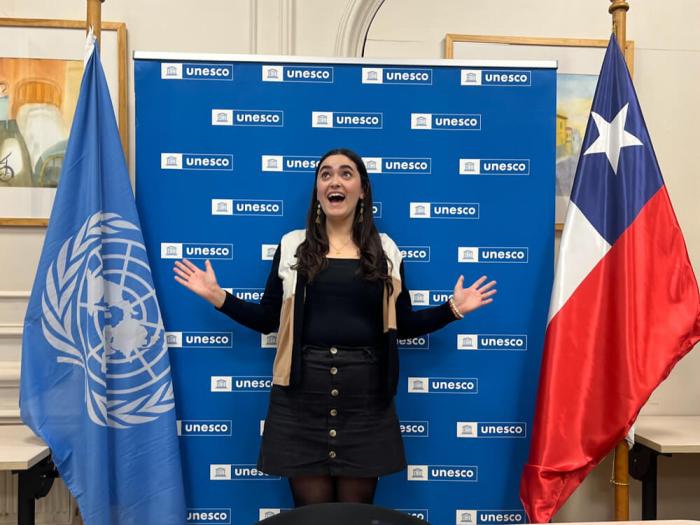 Mia poses for a photo in front of a UNESCO step-and-repeat banner and two flags, excited expression on her face