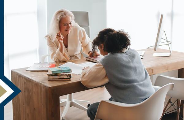 principal sits behind desk talking to middle school student writing on a paper on the desk