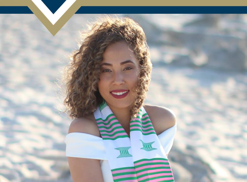 Tatyana Suares headshot wearing graduation stole and seated on a beach with sand in the background