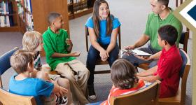 young male school counselor meets with group of elementary students sitting in a circle in the library